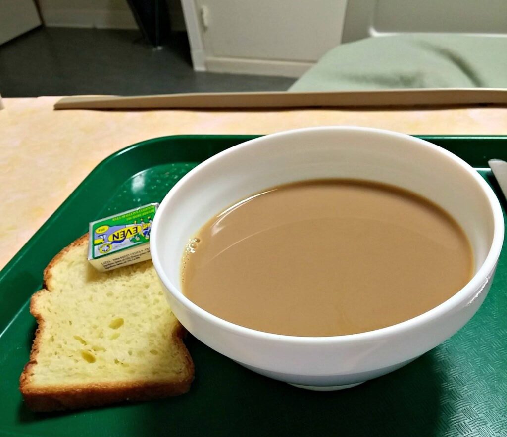 a green hospital tray with a slice of bread on it and a butter packet on that, and a large bowl of what looks to be coffee.