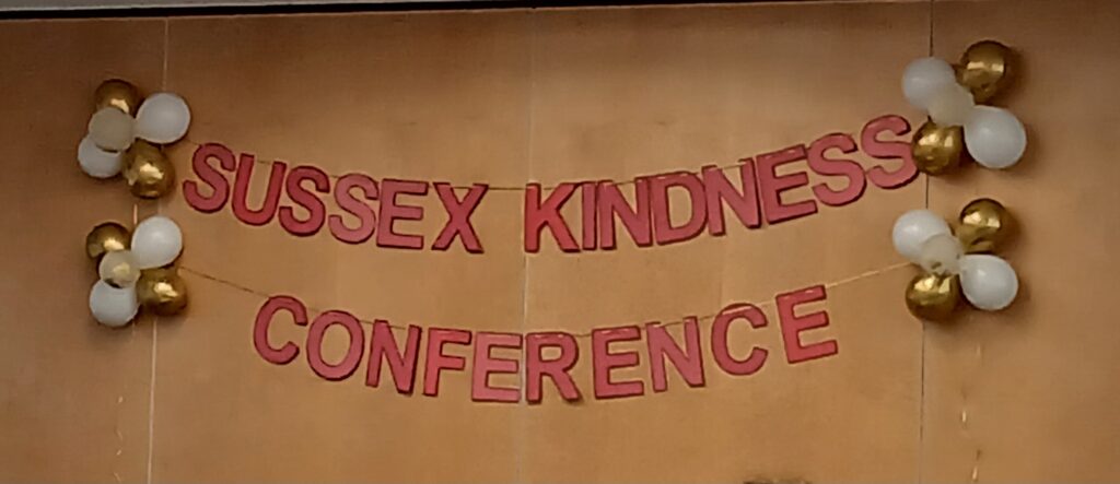 A banner against a wall with white and gold balloons and red letters which read Sussex Kindness Conference