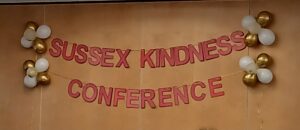 A banner against a wall with white and gold balloons and red letters which read Sussex Kindness Conference