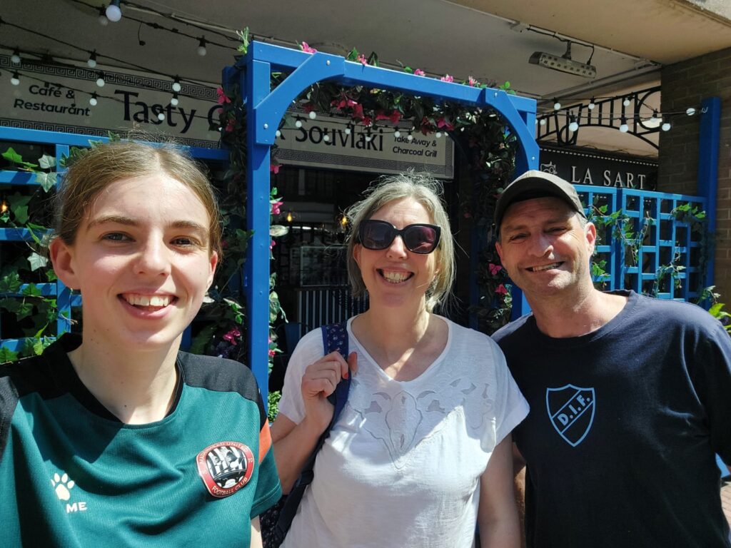 Three people smile outside a restaurant with a blue flower‑covered entrance frame and signs reading “Souvlaki” and “Take Away & Charcoal Grill.”