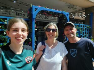 Three people smile outside a restaurant with a blue flower‑covered entrance frame and signs reading “Souvlaki” and “Take Away & Charcoal Grill.”