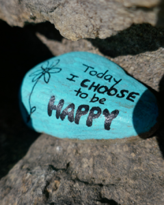 A turquoise painted rock sits among grey stones, decorated with a flower and the handwritten message “Today I choose to be happy.”