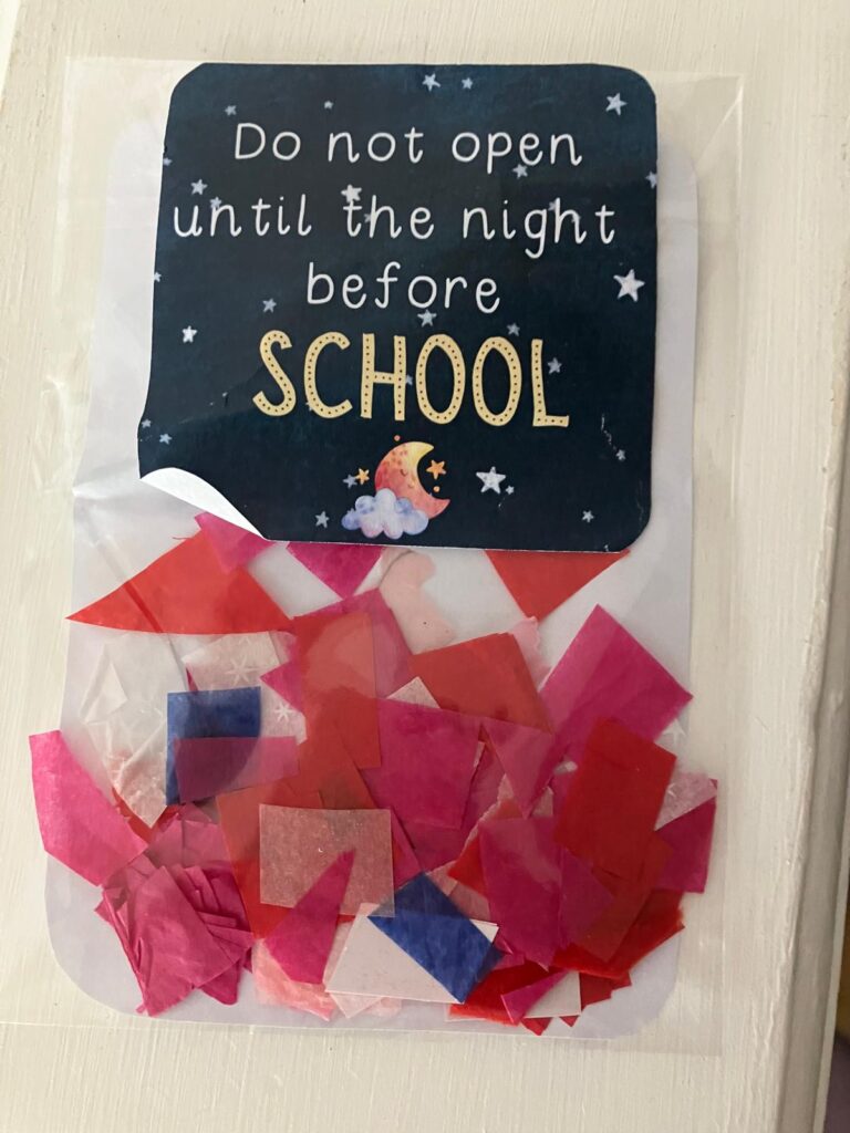 A small plastic bag filled with colourful tissue paper sits on a surface, with a dark blue card attached showing stars, a moon and the message “Do not open until the night before school.”